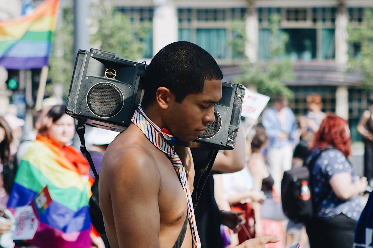 services-09 A shirtless man at a vibrant pride parade carries a boombox on his shoulder, wearing a rainbow necktie.